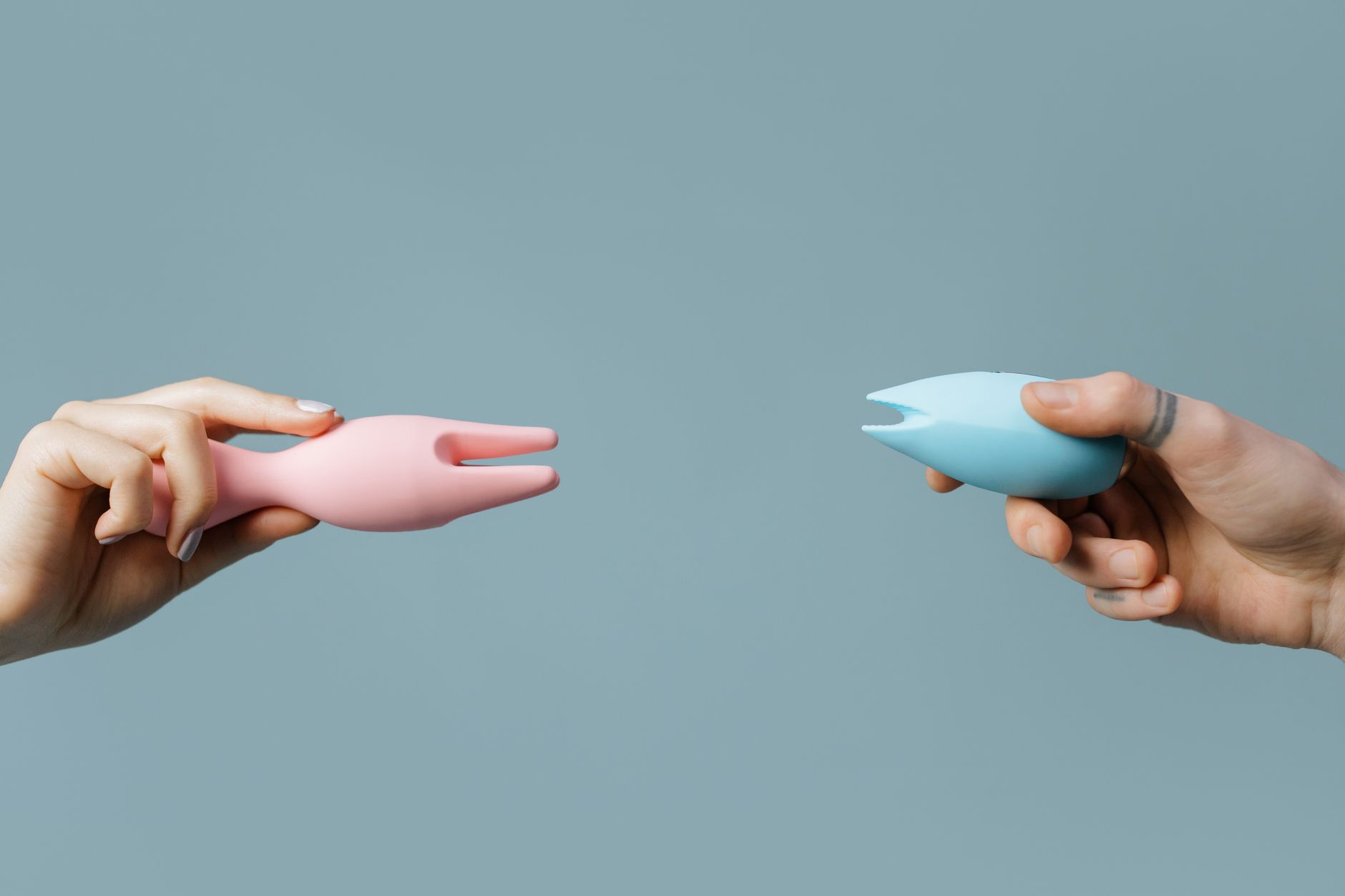 Two hands holding pink and blue vibrators against a pastel background, symbolizing intimacy and partnership.